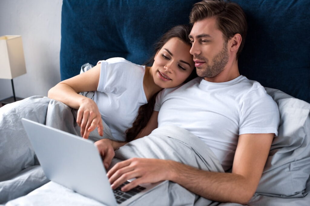 a man and a woman lying in the bad and watching video on the laptop