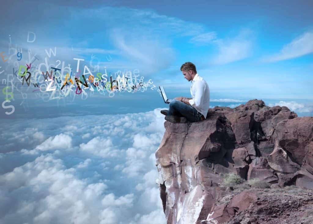 a man sitting on a cliff above the clouds and typing on his laptop